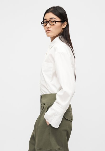 Λευκό μακρυμάνικο πουκάμισο με μια τσέπη στο μπροστινό μέρος, σε συνδυασμό με ψηλόμεσο χακί παντελόνι με πλαϊνές τσέπες και διακοσμητικά μανσαλέτα με τρύπες.