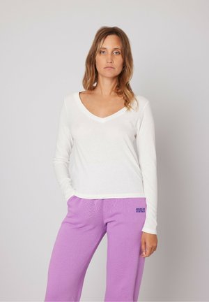 Witte V-hals top met lange mouwen, gecombineerd met paarse sweatpants. Top heeft textuur; de broek heeft een reliëflogo aan de linkerkant.