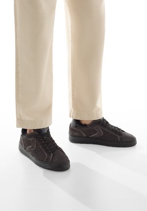 Sneakers in suede marrone con accenti neri, suola piatta e lacci. Indossati con pantaloni beige chiaro, con taglio dritto e fondo a risvoltino.