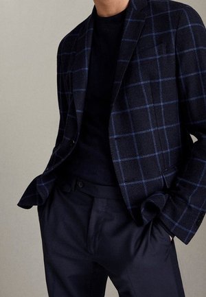 Mężczyzna ubrany w ciemnoniebieską kratkę marynarki na czarnej koszuli z ciemnogranatowymi spodniami, ręce w kieszeniach, na jednolitym tle.