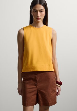 Frau mit langen dunklen Haaren trägt ein ärmelloses gelbes Top, braune Shorts und hält rote und beige Armreifen an ihrem rechten Handgelenk.