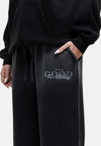 Zwarte hoodie met een voorzak, gecombineerd met zwarte sweatpants. De sweatpants hebben een gedrukt ontwerp met de accenten "GOOD" en "For Nothing".