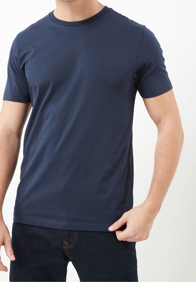 Homme portant un t-shirt bleu marine uni à manches courtes et col rond, ainsi qu'un jean foncé, visible du cou jusqu'aux hanches sur fond blanc.
