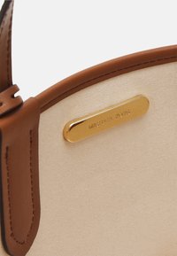 Beige kangasbagi, millel on pruun nahkäär ja rihm, millel on väike kuldplaat, millele on graveeritud "MICHAEL KORS."