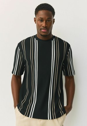 Mann mit kurzen lockigen Haaren, der ein schwarzes kurzärmeliges Hemd mit weißen und beigen vertikalen Streifen trägt, die Hände in den Taschen einer beigen Hose.