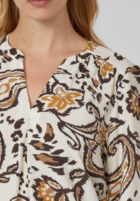 Lichte blouse in crème met complexe bruine en oranje paisleypatronen, met een diepe V-hals en subtiele gerimpelde details op de schouders.