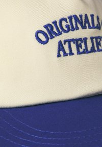 Kasket med en cremefarvet top og blå skygge, der har broderet tekst "ORIGINALS ATELIER" i blå. Materialet er bomuld med synlige stikninger.