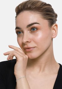 Ritratto ravvicinato di una persona con pelle liscia e chiara e occhi blu. Ha un trucco leggero, capelli castani pettinati all'indietro e un sottile braccialetto d'argento.