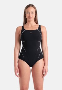 Mulher em pé, de frente, vestindo um fato de banho preto de uma peça com detalhes em branco, contra um fundo claro e simples.
