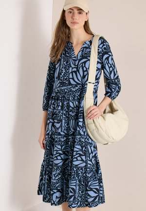 Kobieta w niebiesko-czarnej wzorzystej sukience midi, beżowej czapce i dużej kremowej torbie na ramię, stojąca na tle jednolitej beżowej ściany.