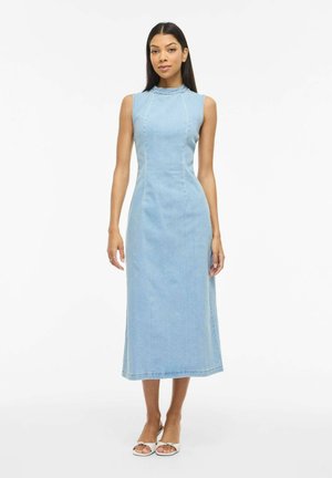 Kvinde stående iført en ærmeløs lyseblå denim midi kjole med høj halsudskæring og hvide åbne tå-hæl på en ensfarvet hvid baggrund.