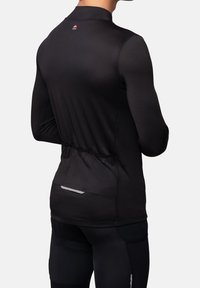 Hombre vistiendo una camiseta de ciclismo negra de manga larga y pantalones cortos con bolsillos traseros y un pequeño logo rojo en la parte superior de la espalda.