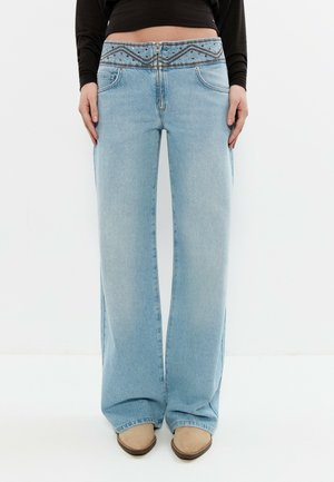 Persona con pantalones vaqueros de pierna ancha de cintura alta en azul claro con cinturilla bordada, camiseta negra de manga larga y zapatos beige puntiagudos.