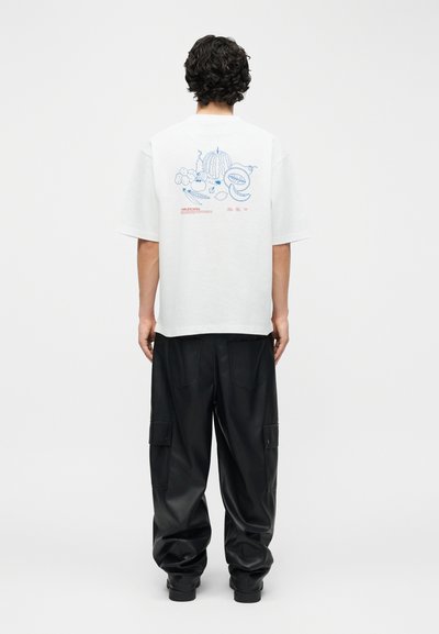 Άτομο με σγουρά σκουρόχρωμα μαλλιά που φοράει λευκό T-shirt με μπλε εικονογραφήσεις φρούτων και κείμενο στην πλάτη, συνδυασμένο με φαρδιά μαύρη παντελόνα cargo.