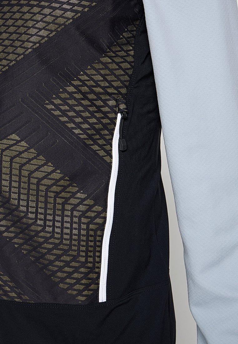 Nahaufnahme einer schwarzen Jacke mit gelbem geometrischem Muster und weißem vertikalen Reißverschluss neben einem hellgrauen Ärmel.