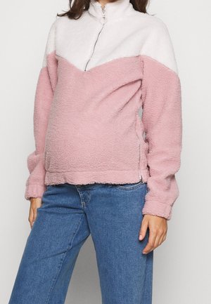 Personne portant un pull en polaire rose et blanc avec une fermeture éclair sur la moitié et un jean bleu, debout devant un fond clair uni.