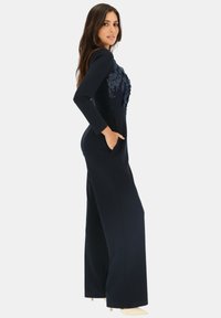 Granatowy kombinezon z długimi rękawami, kwiatowym haftem na korpusie, szerokimi nogawkami oraz bocznymi kieszeniami dla większej funkcjonalności. Gładka faktura materiału.