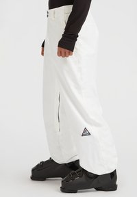 Pantalones de nieve blancos con un corte relajado, cremallera lateral y parche con logo en forma de triángulo. Hechos de material duradero, combinados con botas de esquí negras.