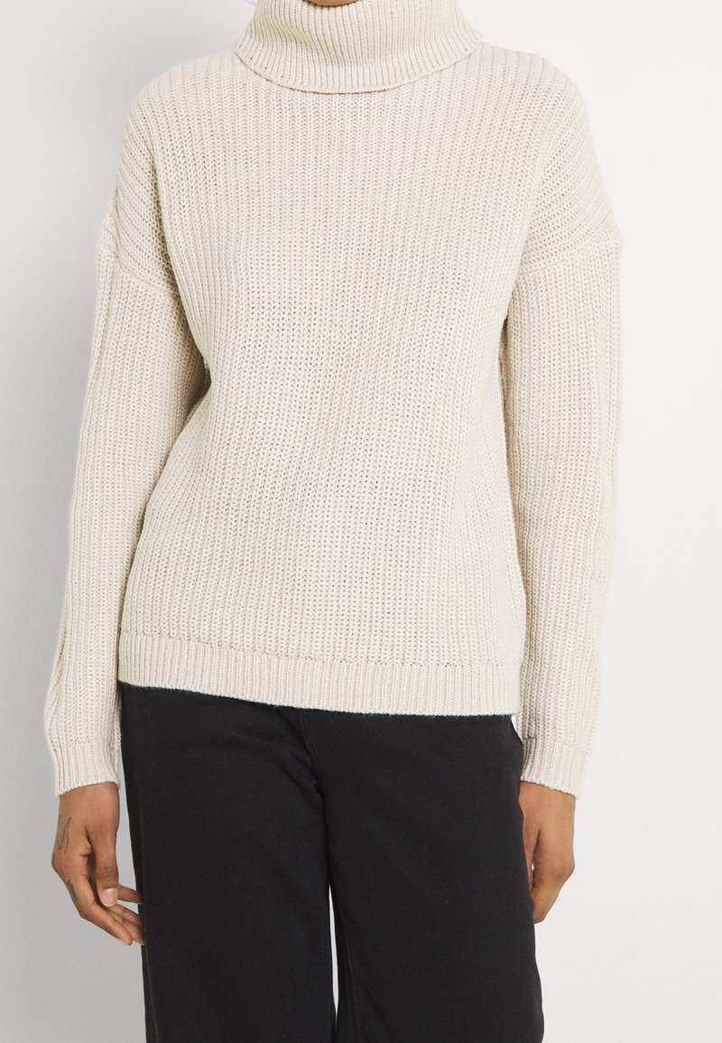 Personne portant un pull à col roulé côtelé de couleur crème à manches longues et un pantalon noir, debout devant un fond uni.