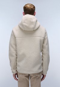 Beige fleece hoodie with a textured surface, full hood, and relaxed fit, paired with light tan trousers. Back view.