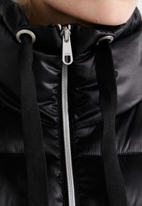 Černá péřová bunda s hladkou, lesklou texturou. Má stříbrný zip, nastavitelné černé šňůrky a vysoký límec.