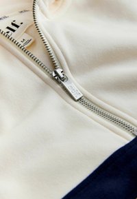 Krémová bunda na zips s tmavomodrým spodkom, vybavená strieborným zipsom a textilnou nálepkou s nápisom "self." Mäkká textúra a ležérny dizajn.