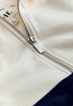 Flødefarvet lynlåstrøje med navy nederst, med en sølv lynlås og et stofmærke med teksten "self." Blød tekstur og afslappet design.