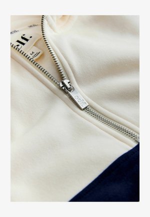 Flødefarvet lynlåstrøje med navy nederst, med en sølv lynlås og et stofmærke med teksten "self." Blød tekstur og afslappet design.