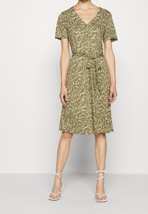 Kvinde iført knælængde olivengrøn blomsterindpakket kjole med korte ærmer og bindebælte, stylet med hvide remsandaler.