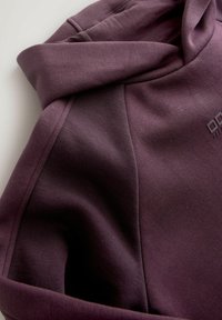 Gros plan d'un sweat à capuche plié couleur prune montrant la texture du tissu, les coutures et une partie de la capuche avec un marquage en relief subtil.