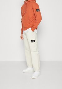 Felpa arancione con tasca frontale, dotata di un'etichetta nera sulla manica. Abbinata a pantaloni cargo beige e sneakers bianche.