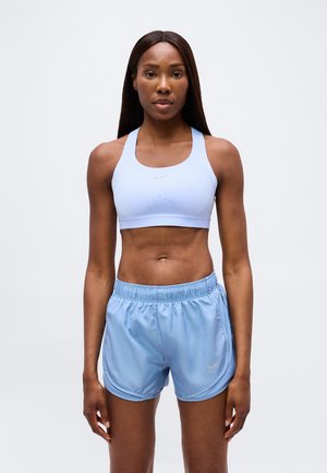 Donna atletica in piedi con il volto rivolto in avanti, indossa un reggiseno sportivo azzurro chiaro e pantaloncini sportivi abbinati su uno sfondo semplice.