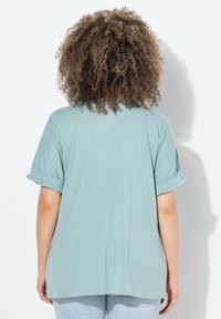 Camiseta de manga corta verde menta con mangas enrolladas, corte relajado y textura suave. La parte trasera presenta un diseño sencillo sin adornos.