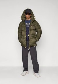 Uomo che indossa una giacca imbottita verde oliva con cappuccio, maglione blu scuro a motivo, pantaloni neri, sneaker bianche, cappellino nero e occhiali da sole scuri.