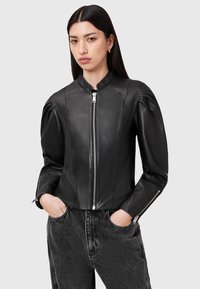 Giacca di pelle nera con un design aderente, colletto alto, maniche a sbuffo uniche e dettagli della cerniera in argento. Abbinata a pantaloni di denim scuro.