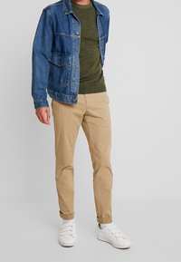 Jeansjacke, olivgrüner Pullover, khakifarbene Hose und weiße Sneakers. Verfügt über Knopfverschlüsse, Taschen und hochgekrempelte Bündchen an den Hosen.