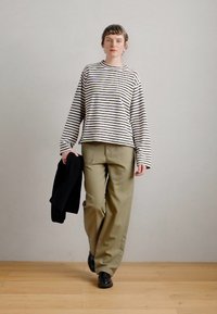 Camisa de manga longa às riscas em creme e azul marinho, combinada com calças largas em verde-oliva. O modelo segura um suéter preto e usa sapatos pretos.