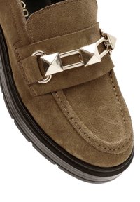 Mocassin en daim marron avec semelle noire et ornements métalliques argentés en forme de pyramide sur la bride avant.
