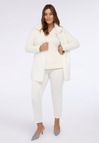 Weißer Cardigan mit einem strukturierten Pelzkragen, kombiniert mit einem weißen Oberteil, das ein dezentes Design aufweist, und weißen Hosen. Das Modell trägt graue spitze Schuhe.