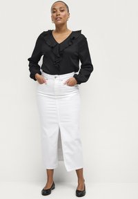 Blusa nera a maniche lunghe con dettagli a volant, abbinata a una gonna di denim bianca con spacco frontale e ballerine nere. Design minimalista.