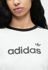White t-shirt with black ribbed neckline, featuring a large black "adidas" logo and a small leaf emblem on the upper left.