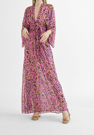 Vrouw die een lange roze jurk draagt met kleurrijke luipaardprint, een diepe V-hals, wijde mouwen, een gestrikte tailleband en gouden sandalen met blokhakken.