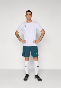 Bílá sportovní košile s krátkým rukávem a černým logem, doplněná tyrkysovými šortkami s číslem a logem, bílými podkolenkami a černými sportovními botami.