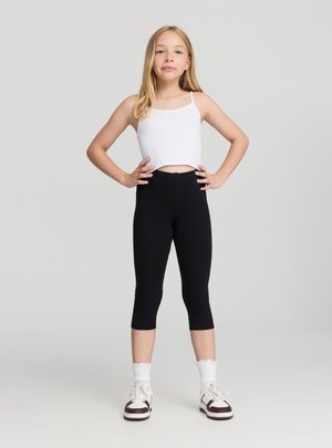 Ragazza giovane in piedi con sicurezza, mani sui fianchi, indossa una canottiera bianca corta, leggings capri neri, calzini bianchi e sneaker su sfondo semplice.