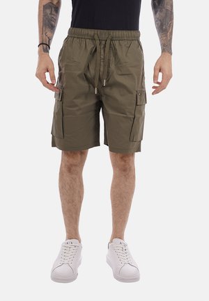Uomo che indossa pantaloncini cargo verde oliva con vita regolabile, maglietta nera, sneakers bianche e tatuaggi sulle braccia visibili dai gomiti in giù.