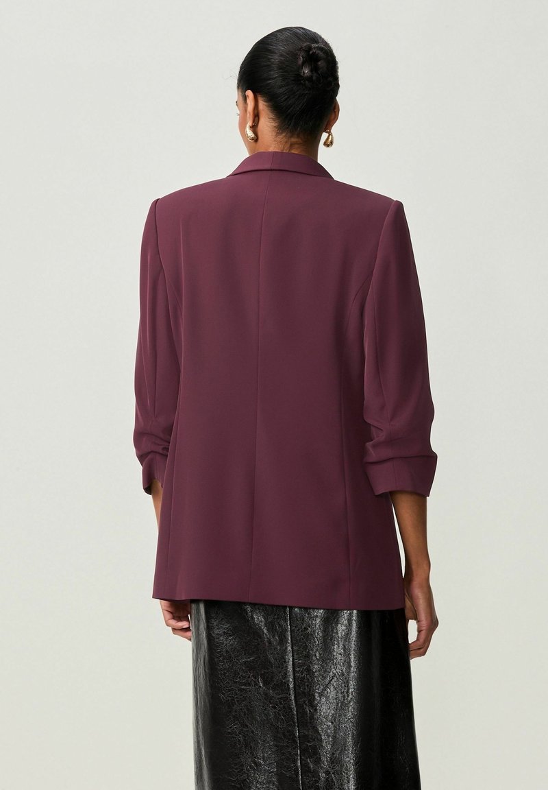 Bordeauxfarbener Blazer mit tailliertem Schnitt, Schulterpolstern und hochgekrempelten Ärmeln, kombiniert mit einem glänzenden schwarzen Rock. Glatte Stofftextur.