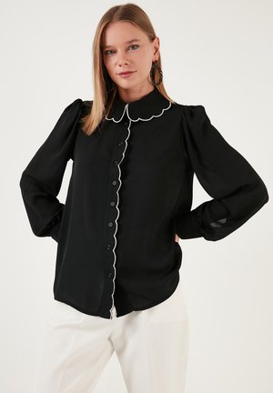 Black blouse with scalloped collar and white trim, button-down front, long puffed sleeves, and lightweight fabric. Worn with white trousers.