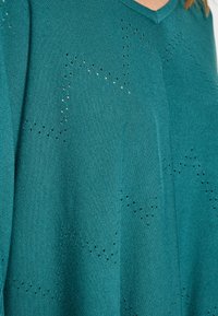 Pulover tricotat de culoare teal cu decolteu în V, prezentând un model texturat format din găuri mici aranjate în forme de stea pe întreaga țesătură.