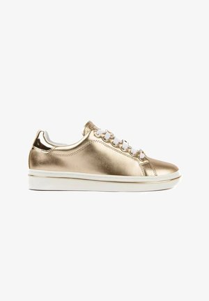 Zapatilla deportiva dorada metálica con suela blanca y cordones blancos, mostrada de perfil sobre un fondo blanco liso.