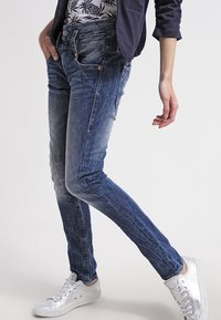 Osoba v vybledlých modrých džínách, květinové košili, tmavě modrém blejzru a stříbrných teniskách, stojící s jednou nohou překříženou.